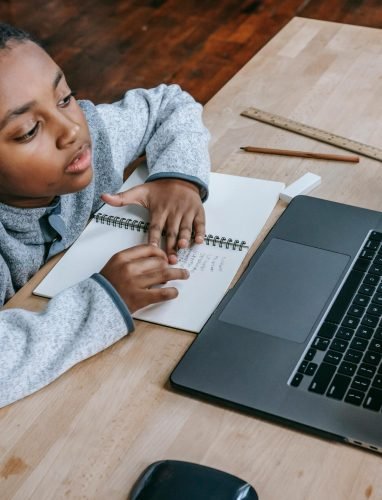 From above of smart cute African American boy studying online with netbook while sitting at table with notebook and pencil